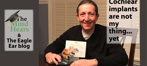 Smiling white man sits at a table with a model of the human ear in front of him. He wears black sweater and behind him are shuttered windows. The text on the right reads " Cochlear implants are not my thing... yet" On the left "the Eagle Ear Blog' is written below The Mind Hears logo.