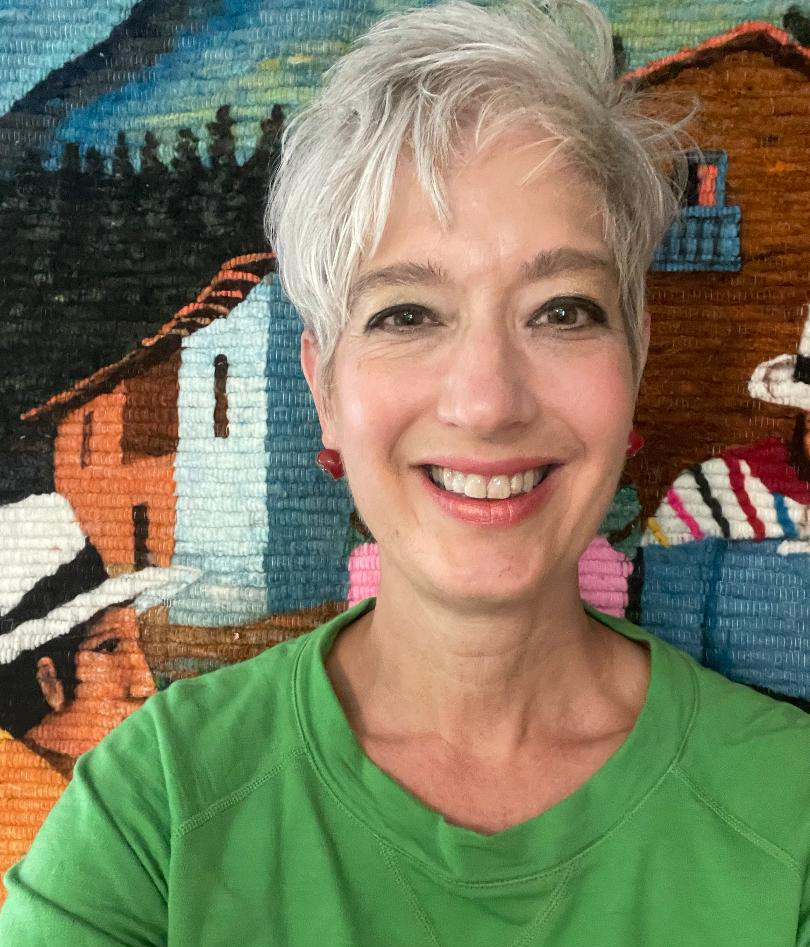 A smiling woman with short white hair, red earrings, and a green top. There is a Latin American tapestry in the background.